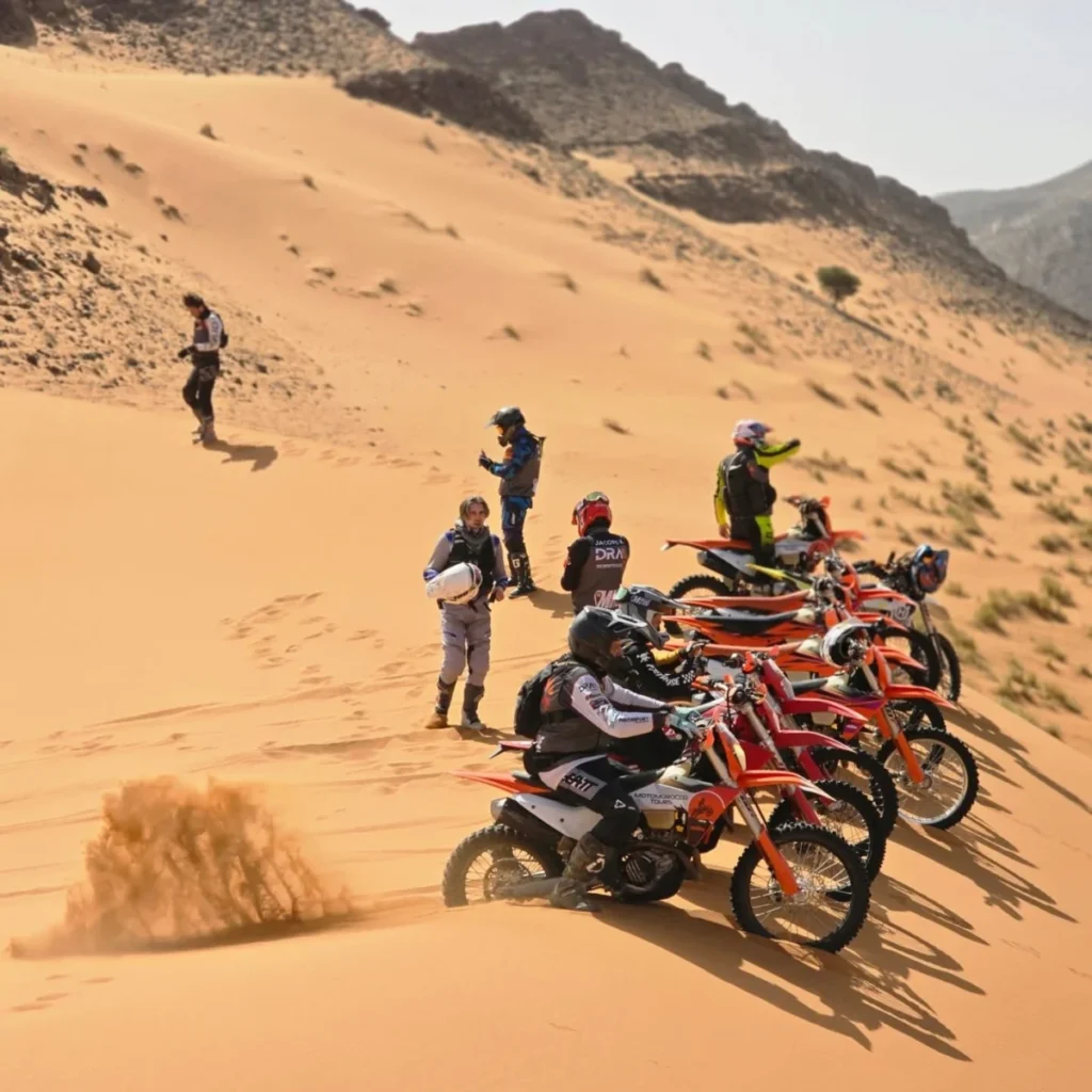 Group of KTM enduro riders lined up on a sand dune below the mountains in Morocco
