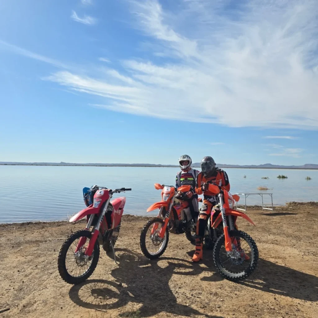 Three riders with KTM enduro bikes beside a desert lake in Morocco